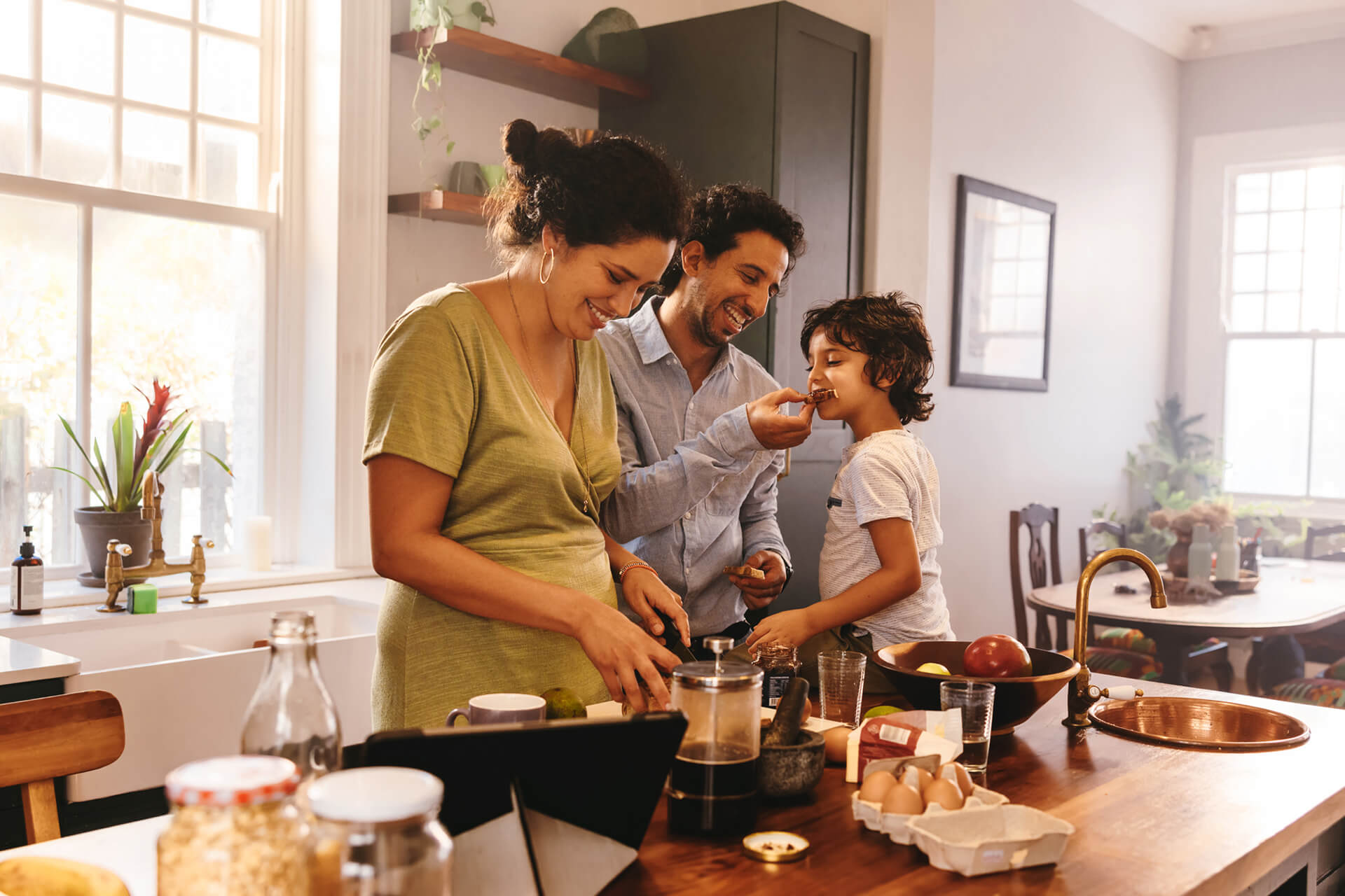 Playful dad feeding his son a slice of bread while his wife prepares breakfast. Family of three having fun together in the kitchen. Mom and dad spending quality time with their son.